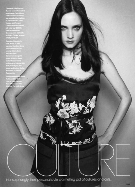 Monochrome image of a brunette woman in an embroidered top and shorts, posing with hands on hips against a neutral backdrop.