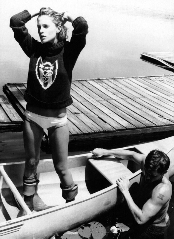 Monochrome shot of a woman standing in a canoe, hands in her hair, and a man at the edge, near a wooden dock and calm water.