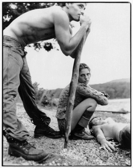 Black and white outdoor shot of a group of three people, two shirtless men and one woman, on a rocky landscape near a lake.