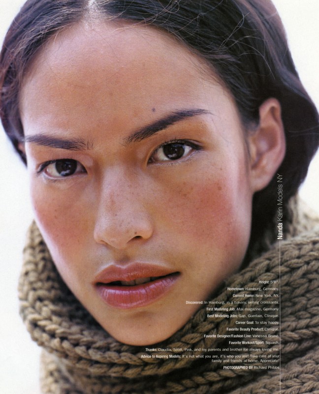 Portrait of an adult female model with dark hair and fair skin, wearing a knitted scarf against a plain white background in a studio setting.