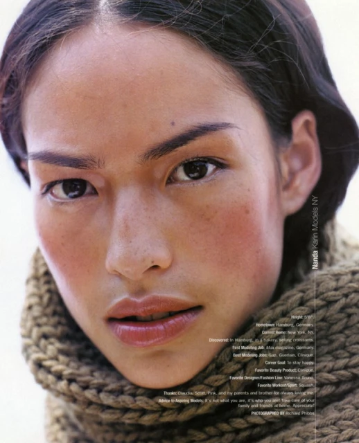 Portrait of an adult female model with dark hair and fair skin, wearing a knitted scarf against a plain white background in a studio setting.