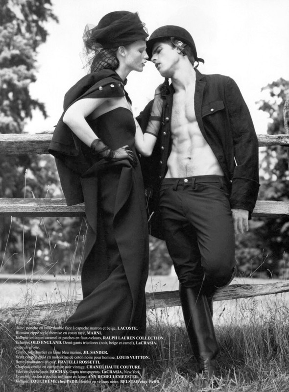 Monochrome fashion shot of a romantic couple near a wooden fence in an outdoor field setting. The woman wears a hat and gloves.