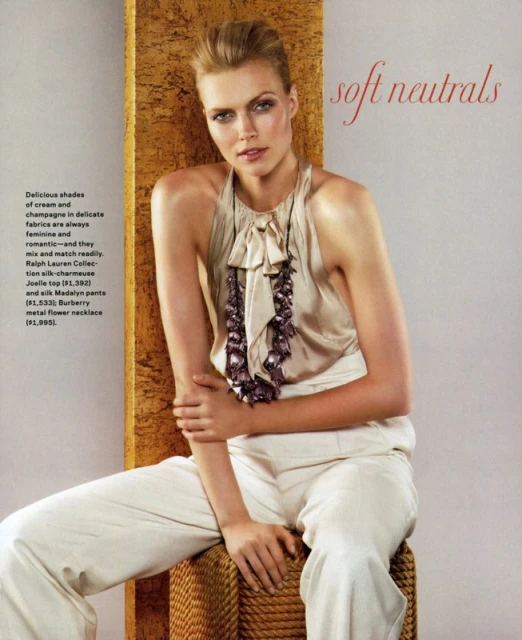 Fashion editorial shot of a woman wearing a beige top and white pants, seated against a textured backdrop in soft, diffused lighting.