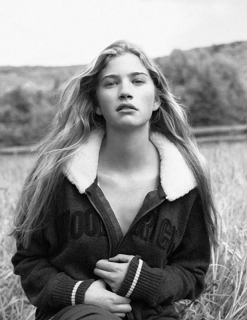 Black-and-white portrait of a blonde woman in a sherpa-collared Woolrich sweater, taken outdoors with a background of blurred grasses and foliage.