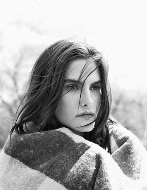 Black and white portrait of a woman with wind-swept hair, wrapped in a patterned shawl, standing outside with trees in the background.