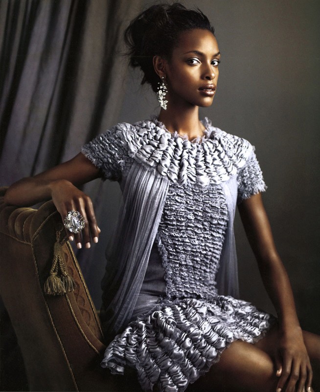 Elegant African American woman poses in a shimmering silver dress, accessorized with chandelier earrings and a statement ring, seated indoors.