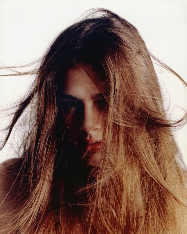 Close-up portrait of a young adult female with long brown hair partially obscuring her face, set against a stark white background.