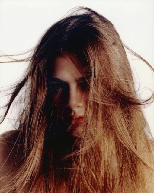 Close-up portrait of a young adult female with long brown hair partially obscuring her face, set against a stark white background.
