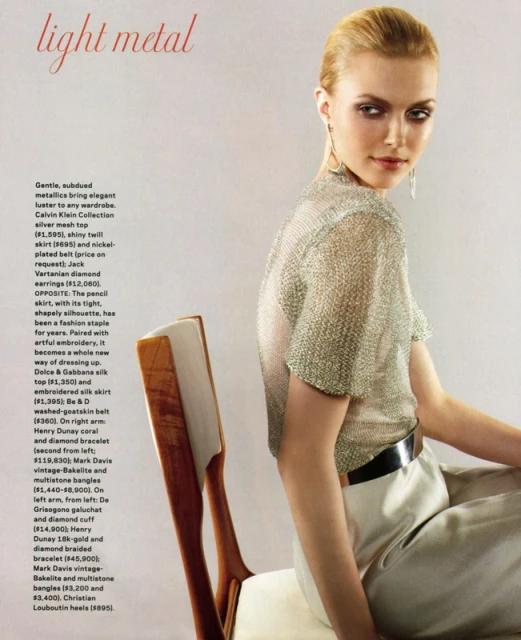 A woman in a metallic top and light pants sits on a chair looking over her shoulder in an indoor fashion setting, editorial.