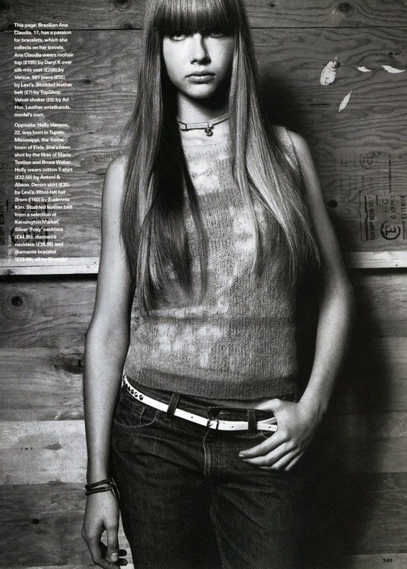 Monochrome editorial portrait of a young woman with long straight hair and bangs wearing a sleeveless top and jeans against a wood wall.