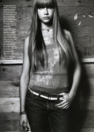 Monochrome editorial portrait of a young woman with long straight hair and bangs wearing a sleeveless top and jeans against a wood wall.
