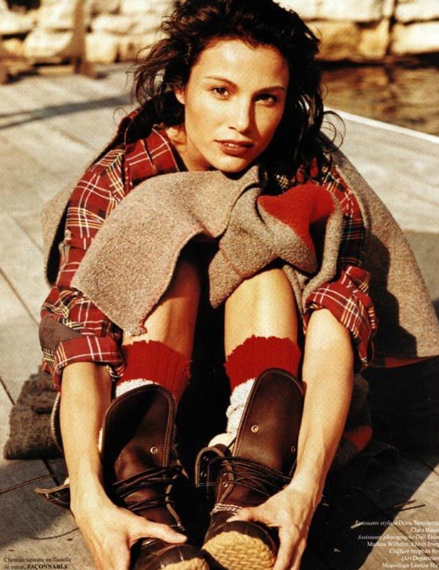 Brunette woman in plaid shirt and boots sitting on wooden pier with a blanket, styled for a fashion editorial shot outdoors.