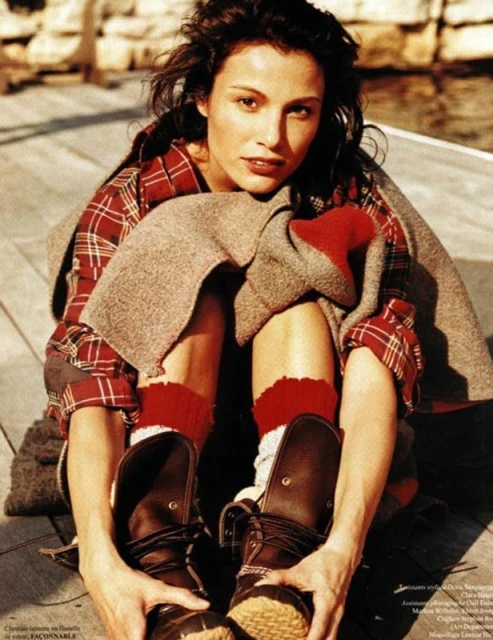 Brunette woman in plaid shirt and boots sitting on wooden pier with a blanket, styled for a fashion editorial shot outdoors.