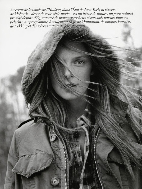 Monochrome portrait of a woman in a hooded jacket, her long hair blowing in the wind, set against an out-of-focus woodland background.