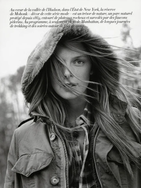 Monochrome portrait of a woman in a hooded jacket, her long hair blowing in the wind, set against an out-of-focus woodland background.