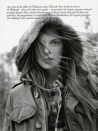 Monochrome portrait of a woman in a hooded jacket, her long hair blowing in the wind, set against an out-of-focus woodland background.