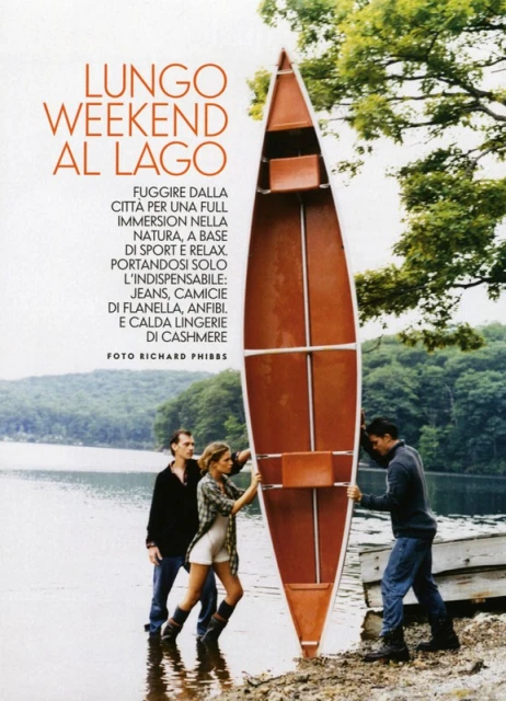 Three people stand on a rocky shore with a brown canoe, a lake and trees in the background, medium shot, fashion editorial.