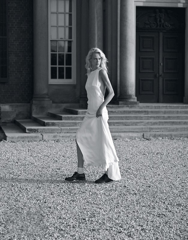 Blonde adult woman in a white dress walks on a gravel path in front of a building with columns and large doors in a black and white outdoor shot.