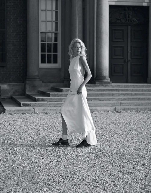 Blonde adult woman in a white dress walks on a gravel path in front of a building with columns and large doors in a black and white outdoor shot.