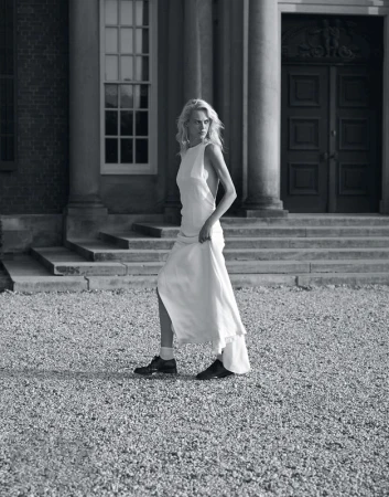 Blonde adult woman in a white dress walks on a gravel path in front of a building with columns and large doors in a black and white outdoor shot.