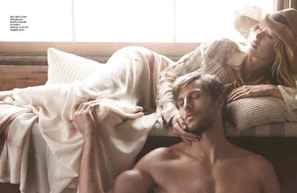 Adult male and female couple relax together indoors; she wears a sunhat and dress while he is shirtless, showcasing lifestyle and intimacy.