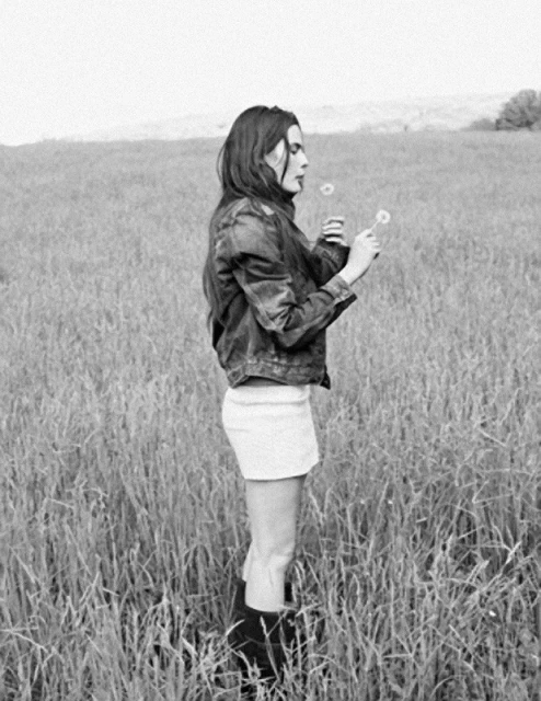 A woman in a denim jacket stands in a field of tall grass holding dandelions, with a light sky in the background, in a black and white photograph.