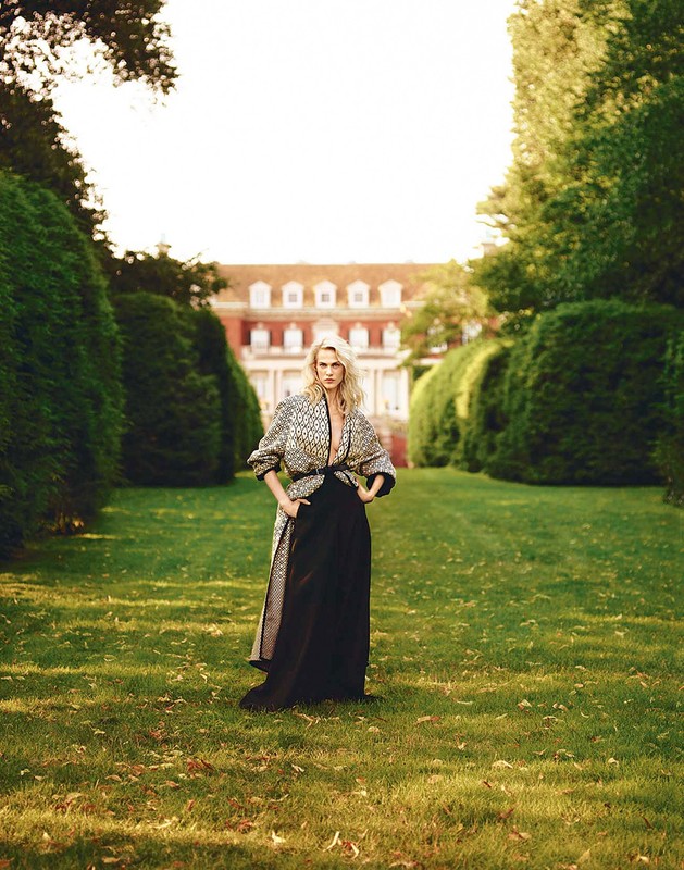 Elegant woman in patterned coat and black skirt stands in a green garden path, backed by trees and a large building in soft daylight.