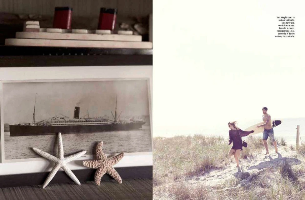 Monochrome photo of a vintage ship alongside a young couple strolling on a sandy beach, holding a surfboard. Beach grasses are visible.