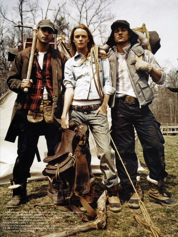 Three outdoor fashion models wearing rugged clothing, including vests, flannel, and denim with boots, standing outside a tent.