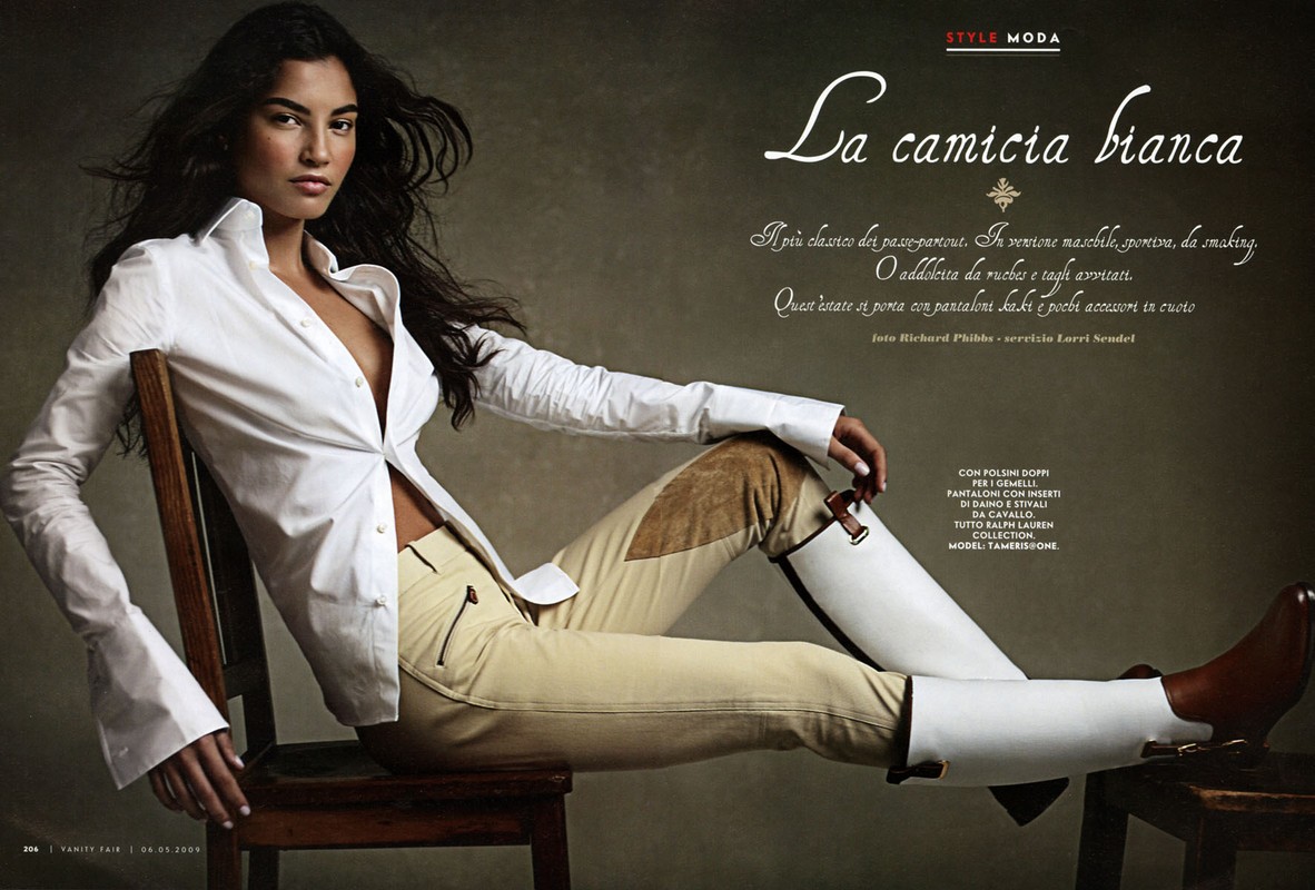 Fashion editorial shot: a slender woman with long dark hair poses sitting on a wooden chair in a white shirt and tan riding pants and boots.