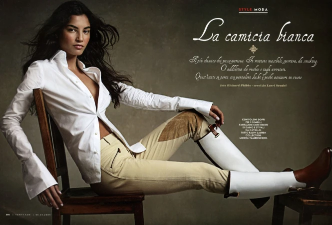 Fashion editorial shot: a slender woman with long dark hair poses sitting on a wooden chair in a white shirt and tan riding pants and boots.
