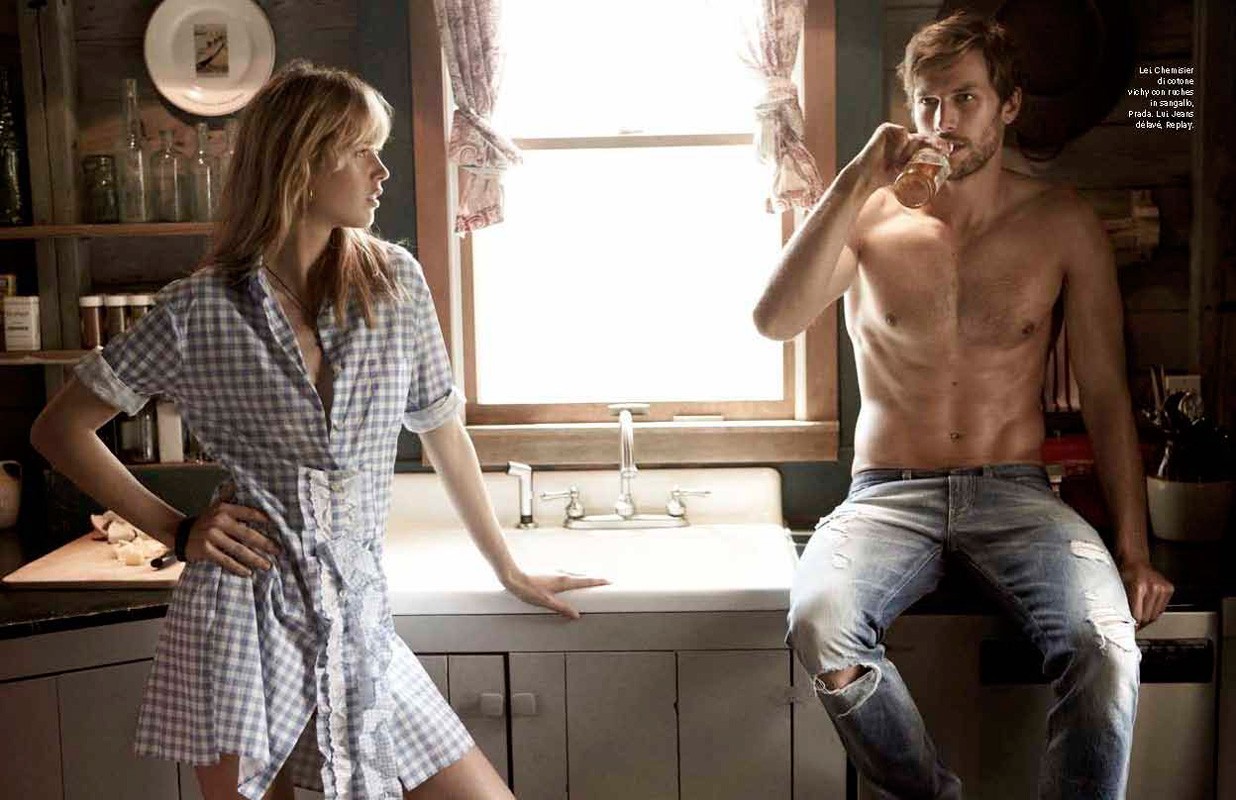 A blonde woman in a checkered dress looks toward a shirtless man holding a glass in a rustic kitchen setting with neutral colors.
