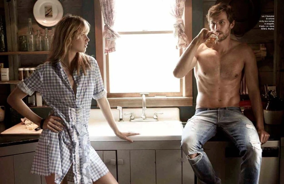 A blonde woman in a checkered dress looks toward a shirtless man holding a glass in a rustic kitchen setting with neutral colors.