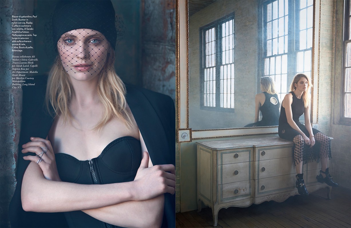 Fashion editorial shot with a blonde woman in a black top and netted head covering, posing near a dresser with a mirror.