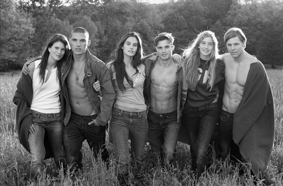 Group of six young adults posing in field, wearing jeans and jackets, photographed in black and white with shallow depth of field.