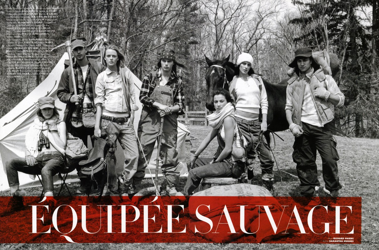 Seven people pose in outdoor fashion editorial with horse, tent, camping gear, and forest backdrop in a black and white photograph.