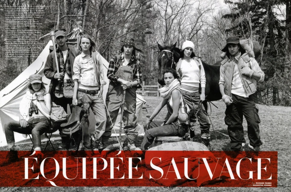 Seven people pose in outdoor fashion editorial with horse, tent, camping gear, and forest backdrop in a black and white photograph.