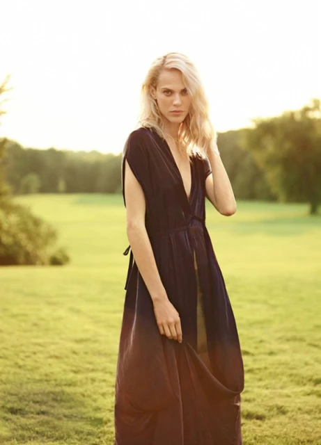 A blonde woman in a dark, low-cut dress stands in a grassy field with a forest backdrop, bathed in golden sunlight, fashion photography.