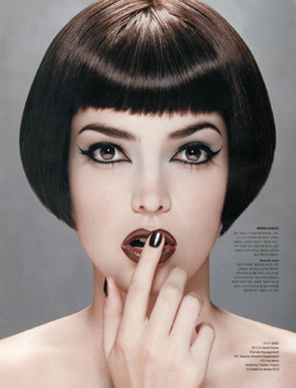 Close-up of a fashionable woman with dark bob haircut, dramatic eye makeup, and glossy brown lipstick, posing with a finger to her lips.