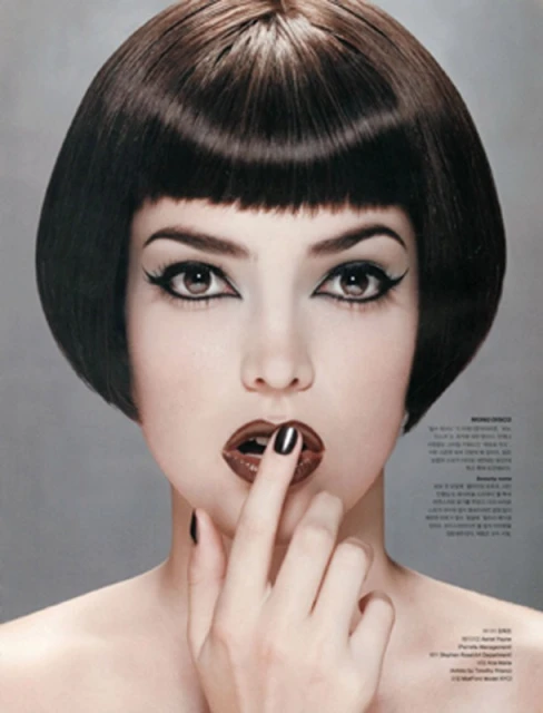 Close-up of a fashionable woman with dark bob haircut, dramatic eye makeup, and glossy brown lipstick, posing with a finger to her lips.