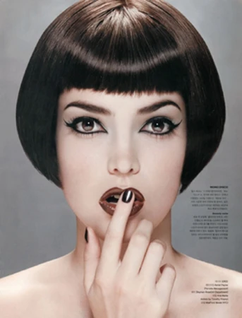 Close-up of a fashionable woman with dark bob haircut, dramatic eye makeup, and glossy brown lipstick, posing with a finger to her lips.