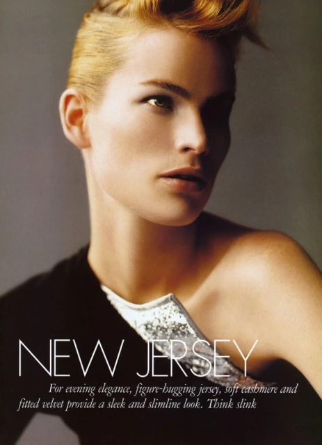 Close-up fashion shot featuring a blonde adult woman with short hair and a black top with a silver detail against a grey background.