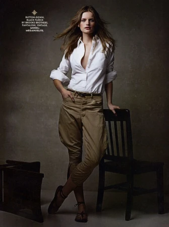Fashion editorial shot of a woman wearing an unbuttoned white shirt and jodhpurs, leaning against a chair in a photo studio.