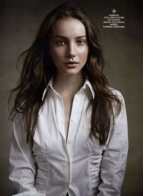 Portrait of a young woman with long brown hair, wearing a white button-down shirt, against a neutral-toned background, fashion editorial.