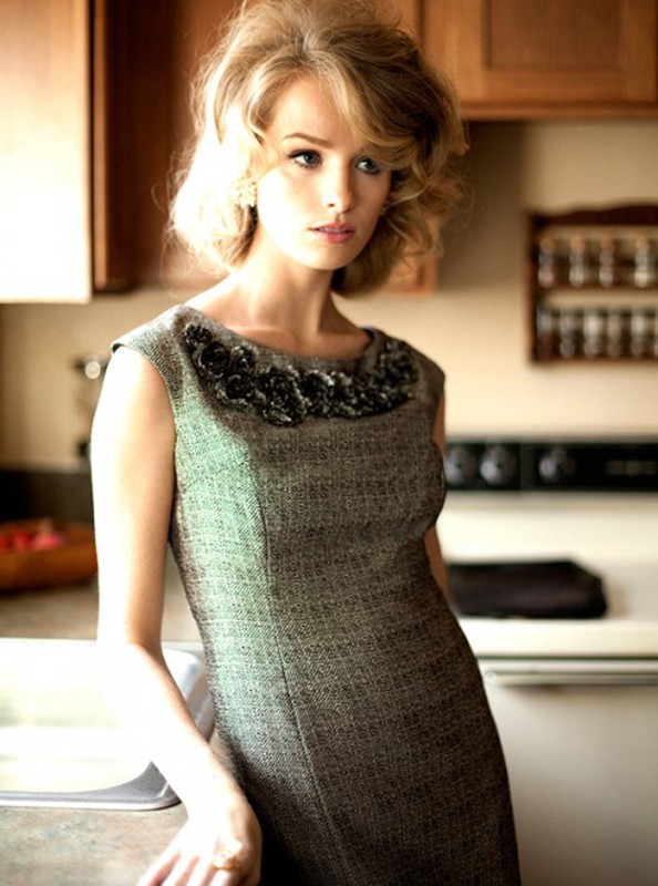 Woman in gray dress with ruffled neckline standing in a kitchen. She has blond hair and is looking off to the left.