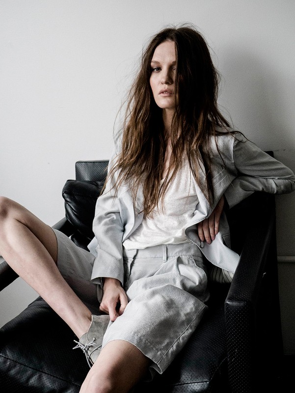 Adult woman in a gray suit jacket and shorts sits in a black leather chair in an indoor setting with neutral-colored walls.