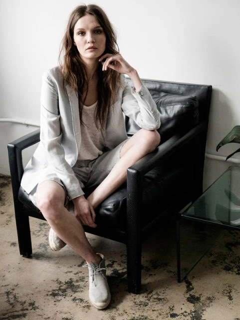 A young adult woman in a blazer and shorts is seated in a black chair, looking at the camera with a neutral expression in an indoor setting.