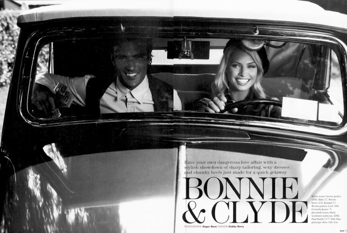 Black and white photograph of a smiling couple in a vintage car, woman driving, set against an out-of-focus greenery backdrop.