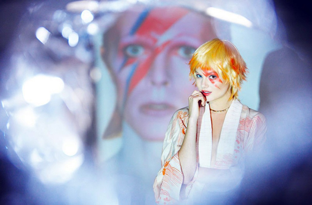Woman with orange hair and face paint, wearing kimono, poses in front of a blurred portrait of a man with lightning bolt face paint.