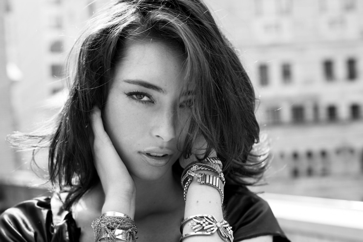 Monochrome portrait of a woman with dark hair, wearing bracelets, hand near her face, and a blurred cityscape in the background.